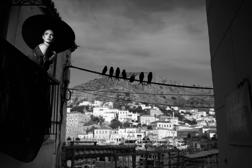 Ședință foto în Grecia, Dark Bride pe insula Hydra: mireasa în negru, cu pălărie cu boruri largi, privind spre port, cu păsări pe un fir