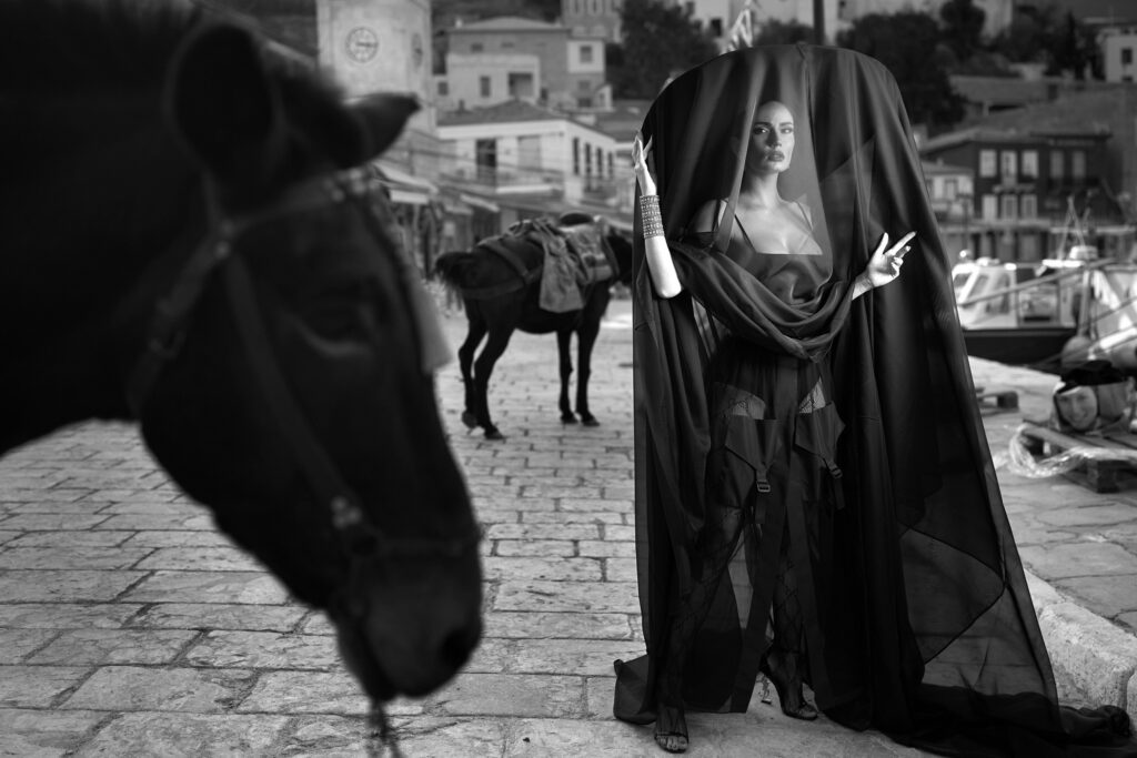 Mireasa în negru pe cheiul din Hydra, lângă un măgar, cu casele insulei în fundal