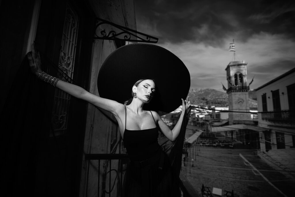 Dark bride sprijinită de un stâlp de fier forjat pe o terasă din Hydra, cu turnul mănăstirii în depărtare