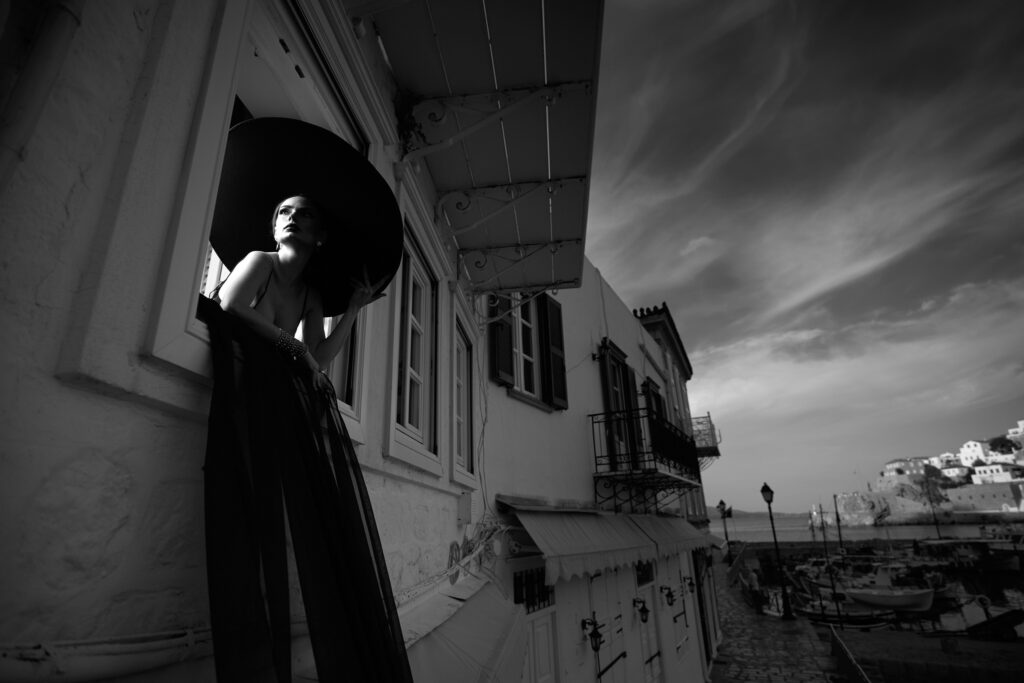 Mireasa în negru la un balcon alb din Hydra, cu voalul atârnând și cerul cu nori în mișcare
