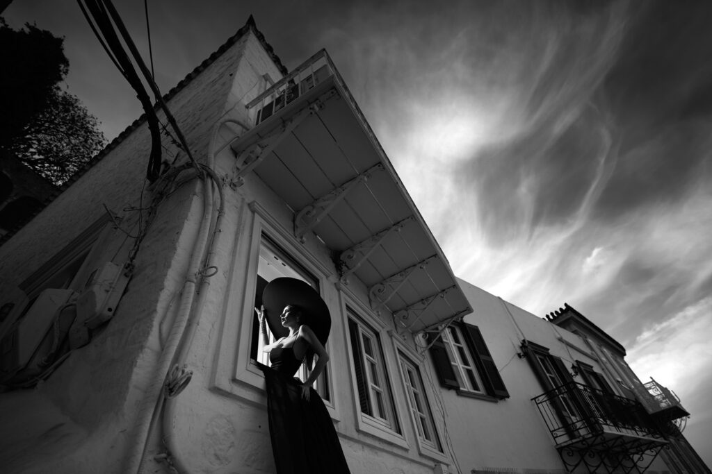 Casă albă din Hydra fotografiată de jos, cu mireasa în negru la fereastră și cer dramatic