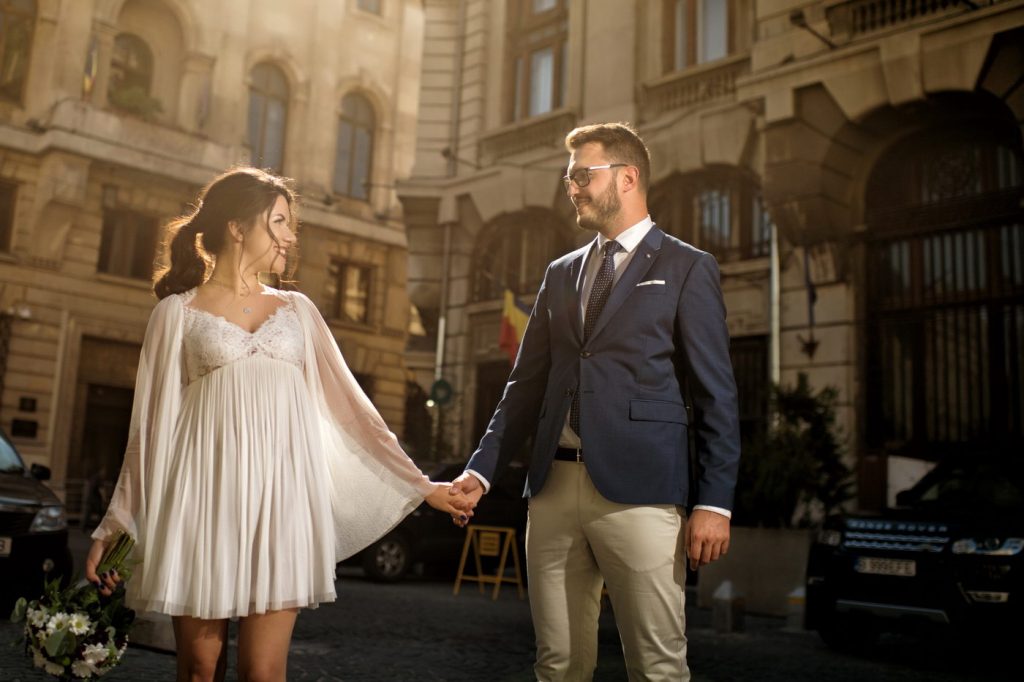 Mirii se țin de mână pe o stradă cu clădiri vechi, mireasa în rochie scurtă