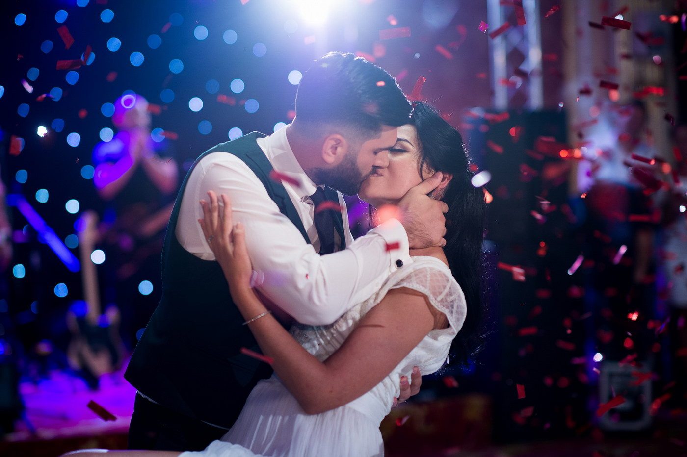 Mirii se sărută în timpul dansului mirilor, cu confetti roșii și lumini albastre în sală