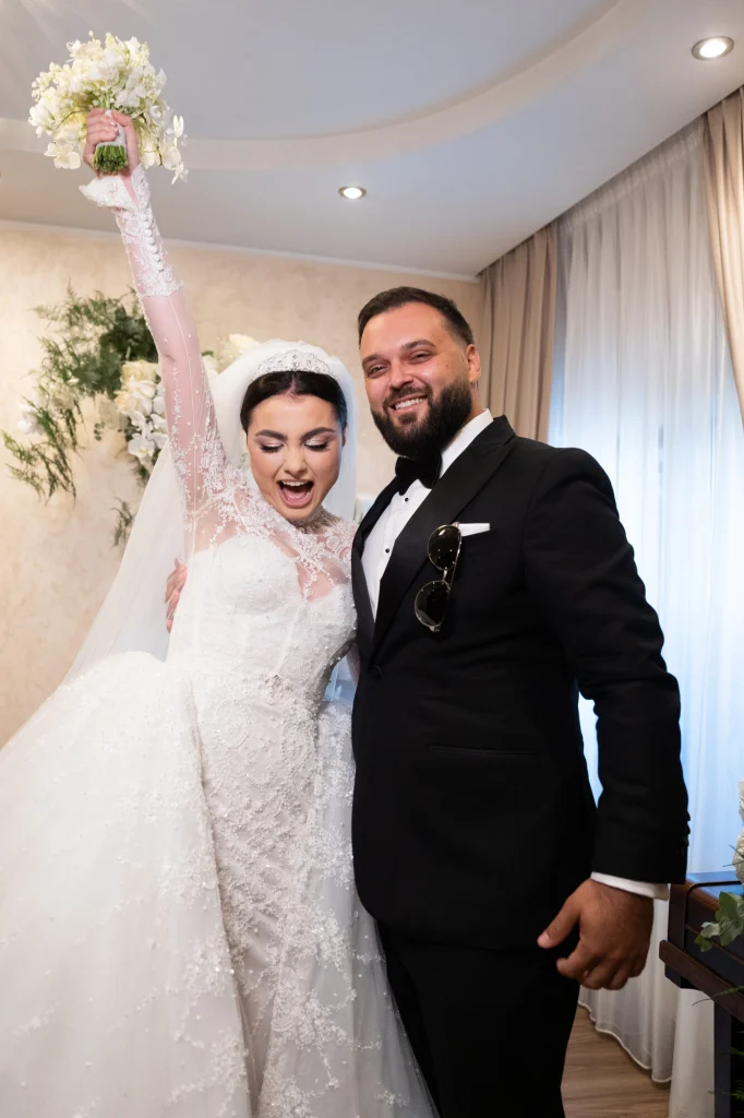 Mireasa ridică buchetul deasupra capului și râde lângă mire