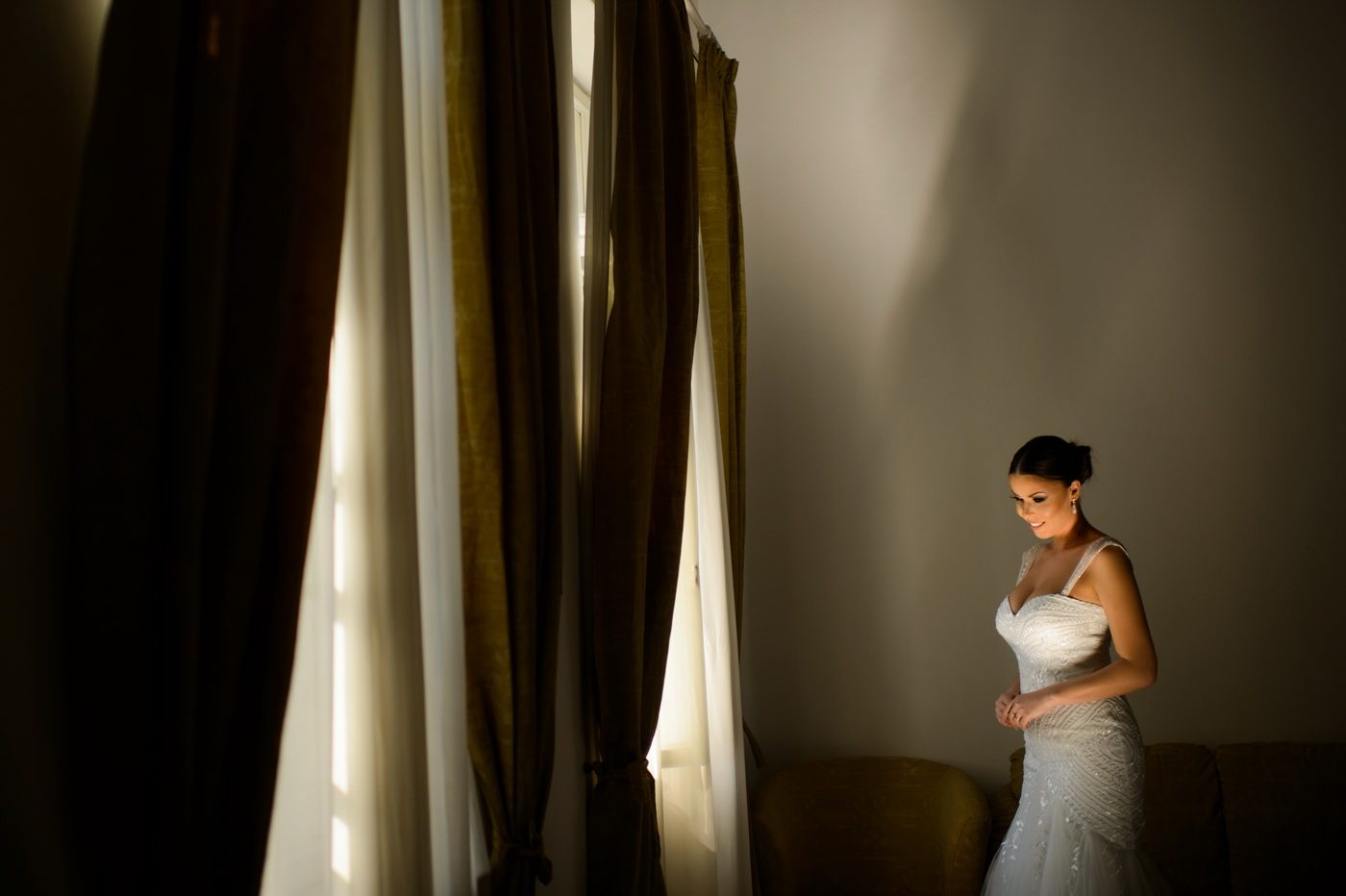 Mireasa în rochie albă stă lângă o fereastră cu draperii grele, luminată cald, într-o cameră în penumbră