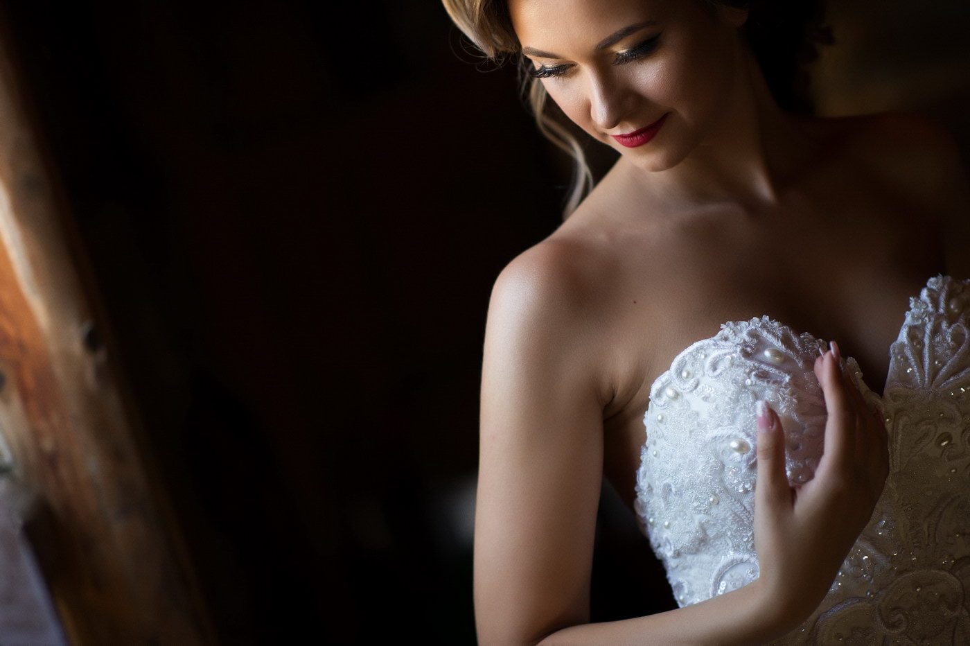 Mireasa zâmbind cu privirea în jos, cu mâna pe corsajul de dantelă al rochiei, luminată de la fereastră
