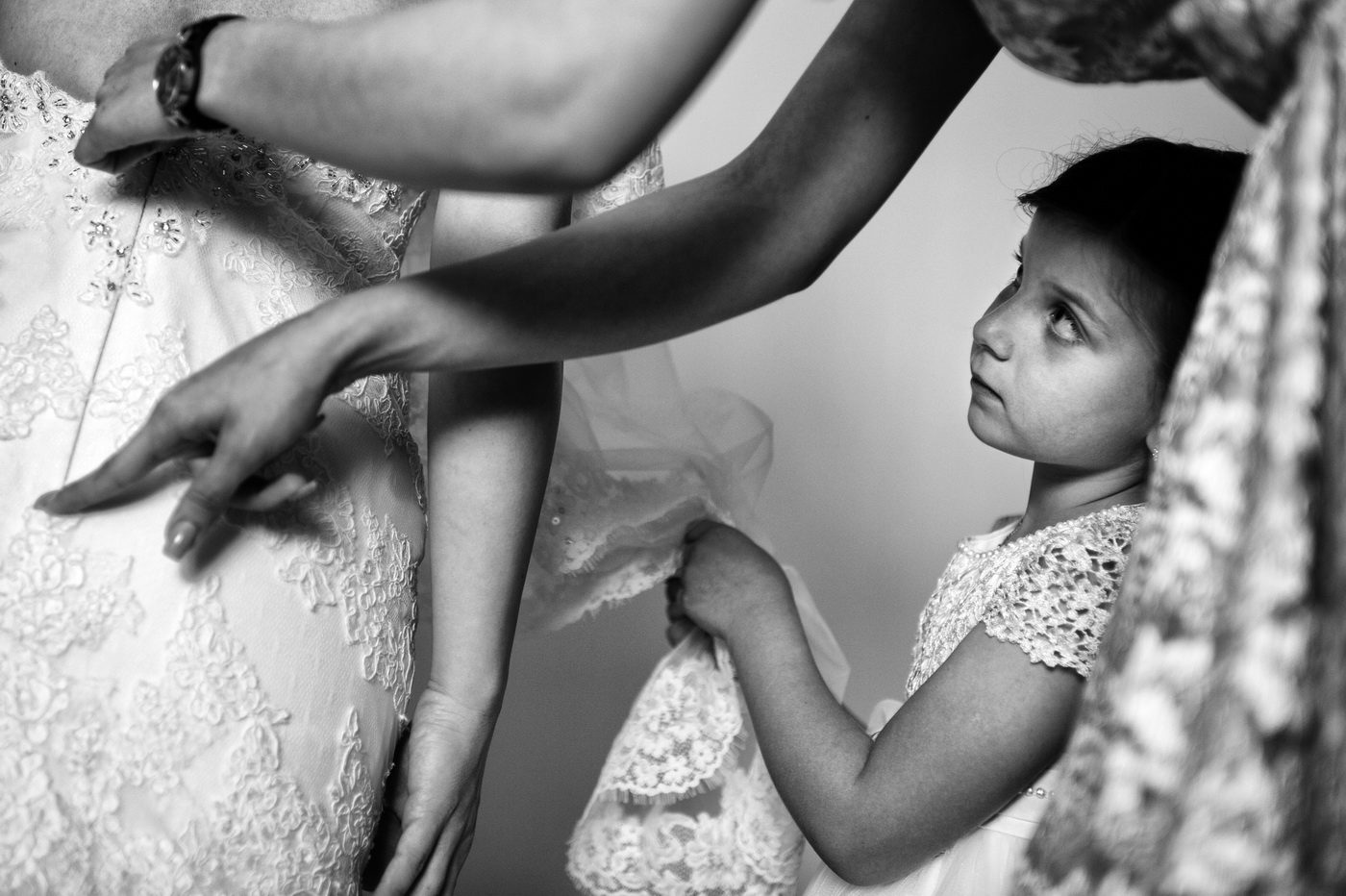 Pregătiri de nuntă: o fetiță privește în sus cum se încheie rochia miresei, fotografie alb-negru